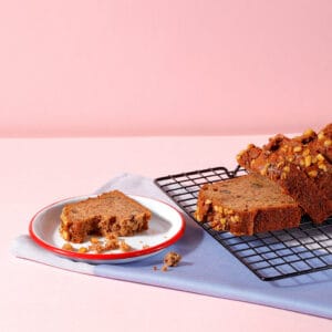 Banana and Walnut Loaf slices on a cooling rack with a slice and crumbs on a red rimmed plate.
