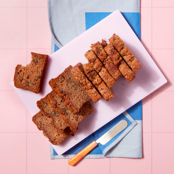 A whole tray of Banana and Walnut Loaf cake on a pink chopping board sitting on a blue and grey napkin.
