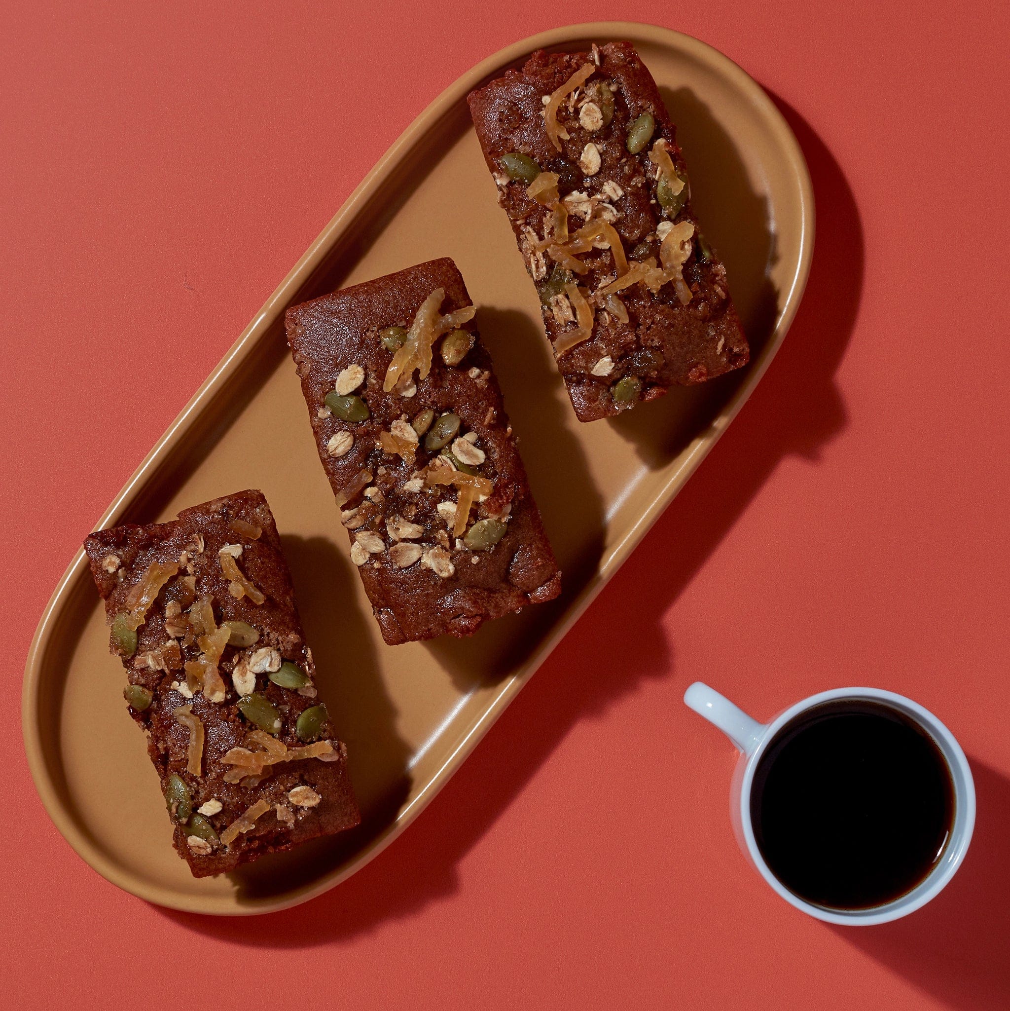 Vegan Sticky Ginger and Lemon Mini Loaf topped with lemon zest and seeds beside white mug for café display.