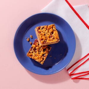 Vegan Berry Crumble slices with golden oat topping and jam layer on blue plate – plant-based coffee shop bake.