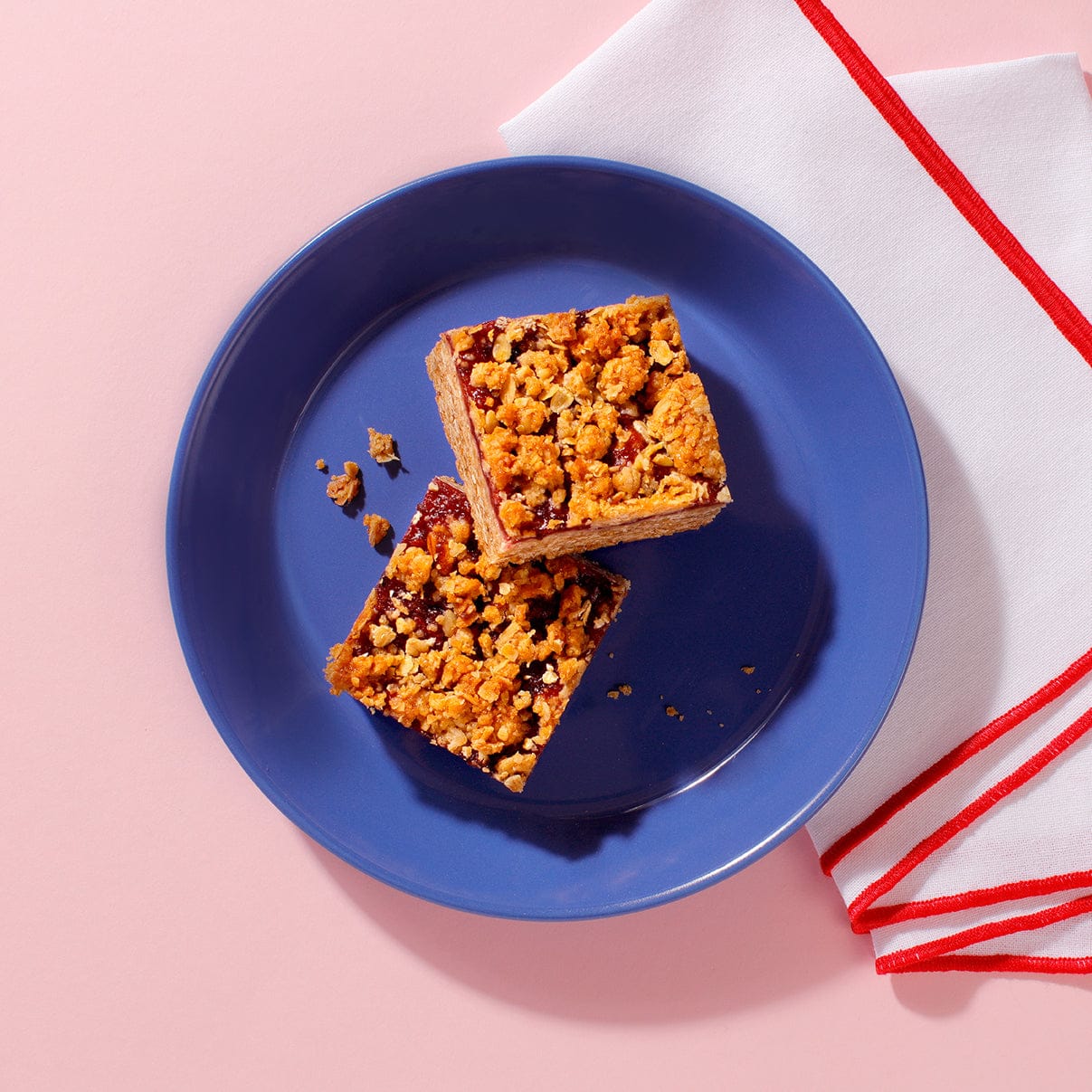 Vegan Berry Crumble slices with golden oat topping and jam layer on blue plate – plant-based coffee shop bake.