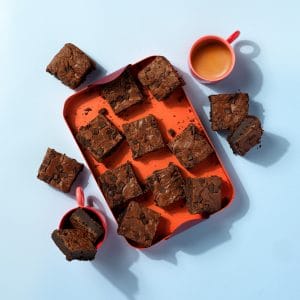 Slices of gluten free Choc Chunk Brownies on an orange tray next to a coffee cup on a light blue background.
