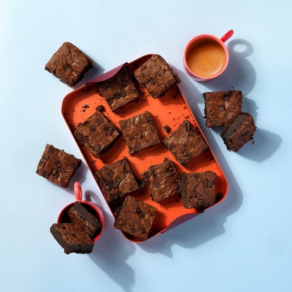 Slices of gluten free Choc Chunk Brownies on an orange tray next to a coffee cup on a light blue background.