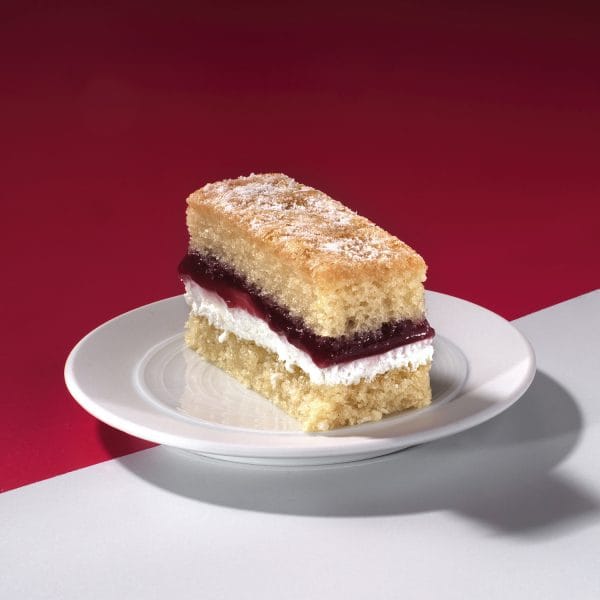 Close-up of Victoria Sponge Traycake slice with jam and cream layers on white surface.