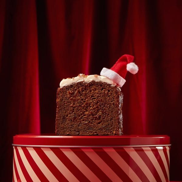 A single slice of rum and raisin Christmas loaf cake standing on a red striped tin against a red curtain backdrop.